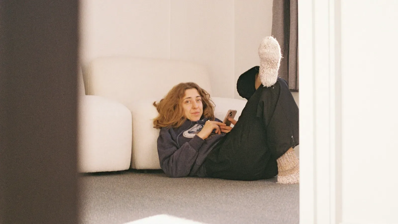 A woman lying on the floor of a bright living room, propped against a couch with her feet up, looking at her phone.