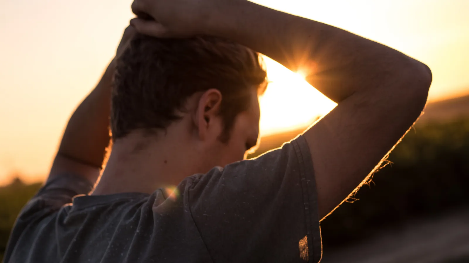 A man with both hands on his head, silhouetted against a low evening sun.