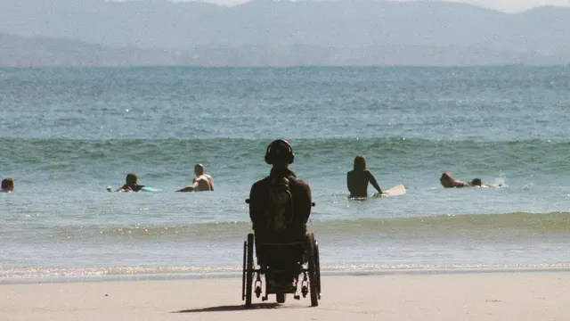 A person in a wheelchair on the sand, facing the sea and watching surfers in the water.