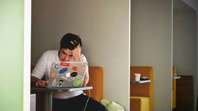 A man at a laptop in a small booth, holding his head in his hand.