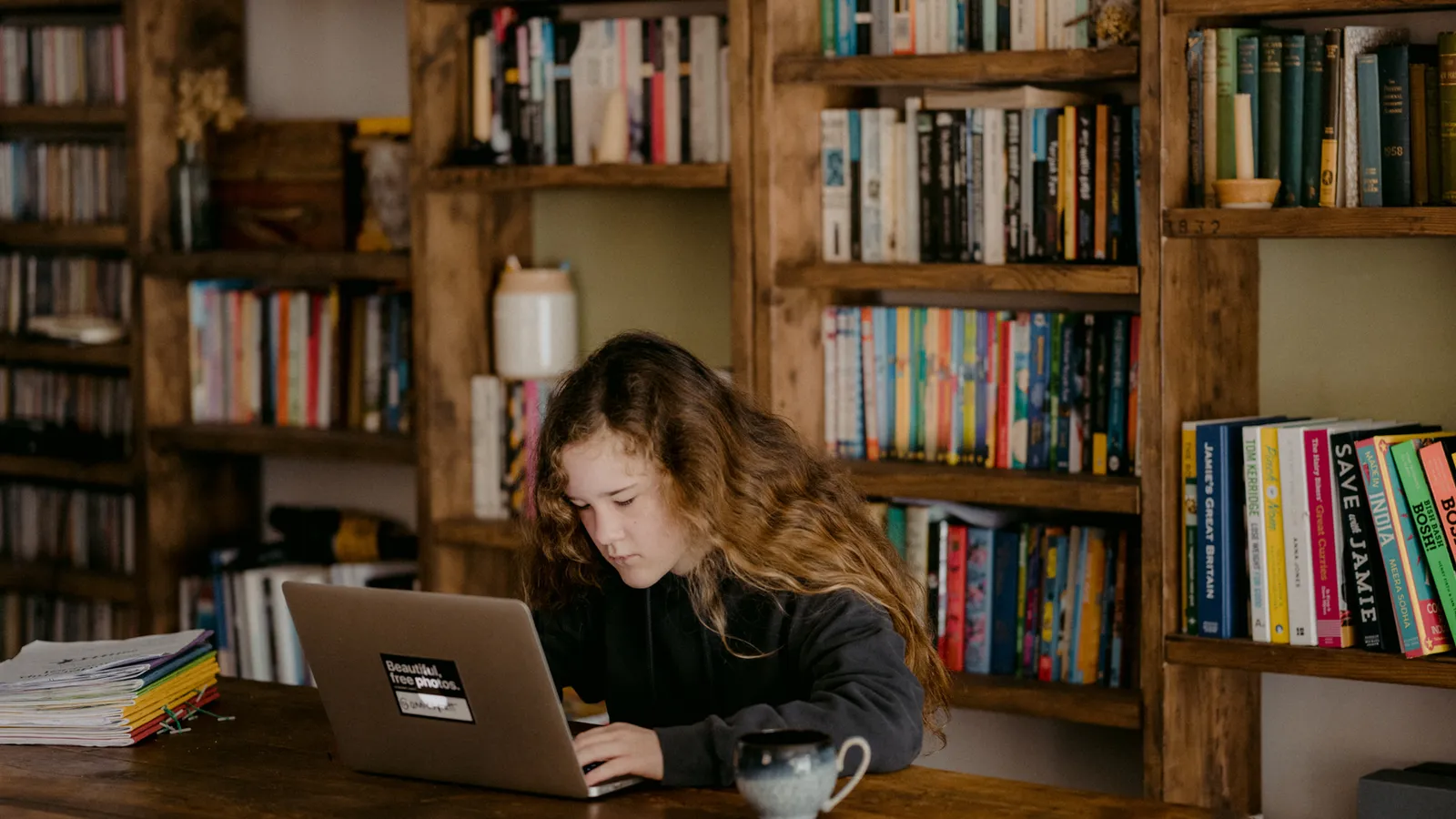 A young person working on a laptop at a table in front of full bookshelves.
