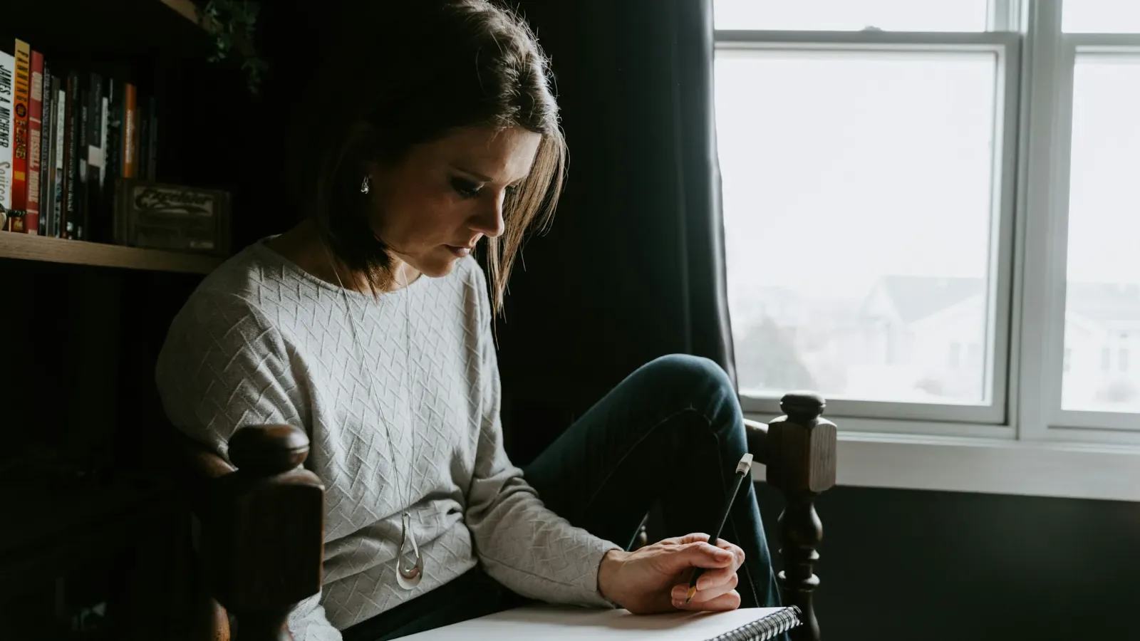 A woman sitting in a chair by a window, writing in a notebook on her lap.