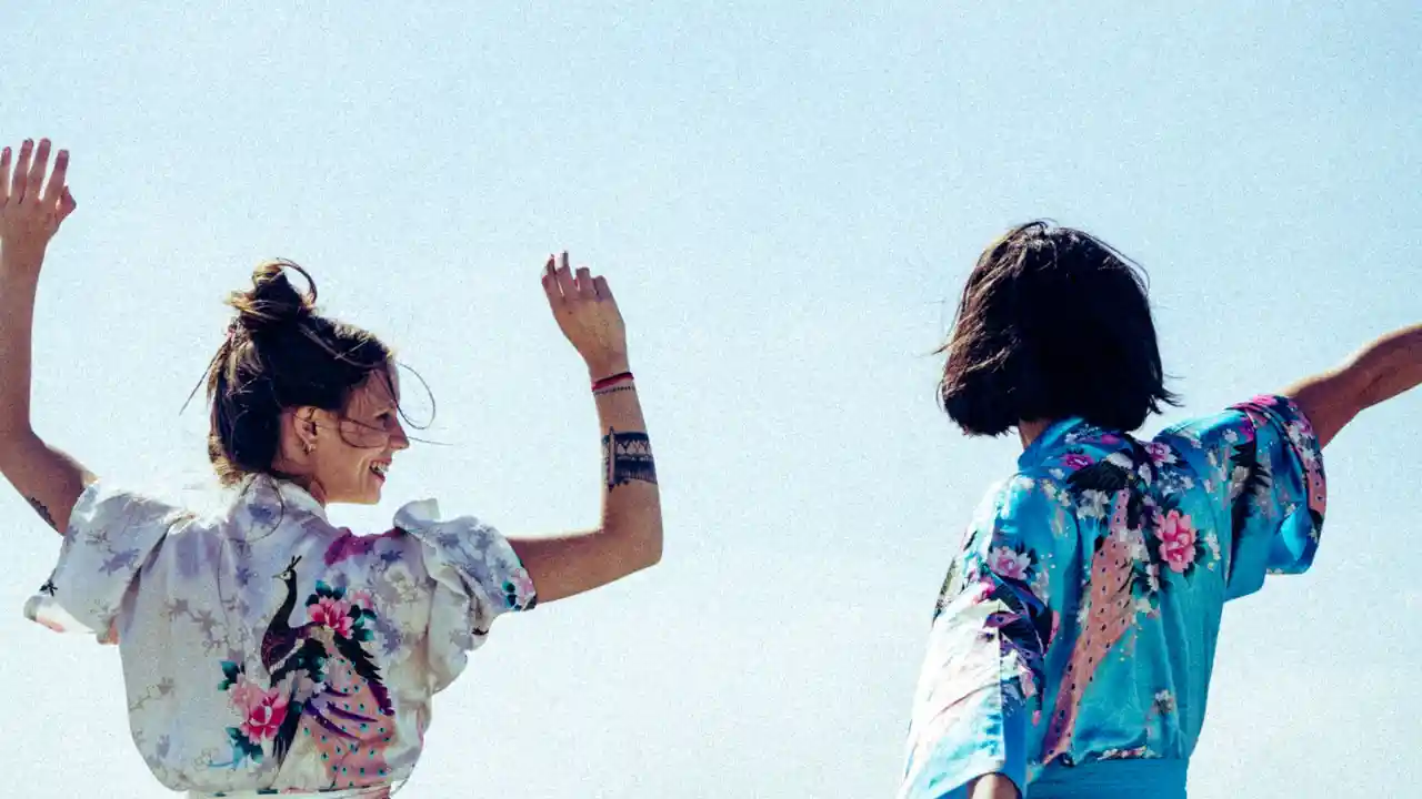 Two young women in bright kimono-style shirts, arms raised against a clear blue sky.