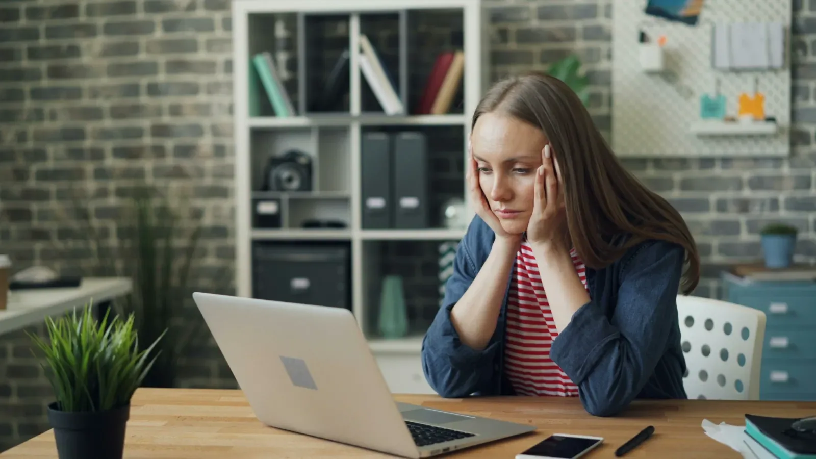 A woman at a desk resting her head in her hands, looking at her laptop.