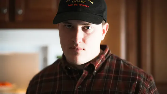 A young man in a cap and flannel shirt looking steadily at the camera.