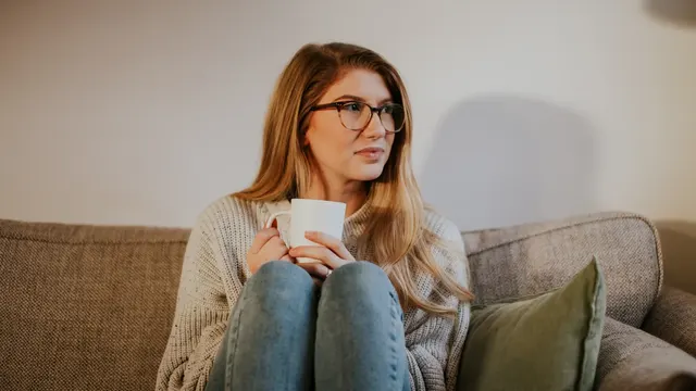 A woman in glasses sitting on a couch with a mug, looking out of frame.