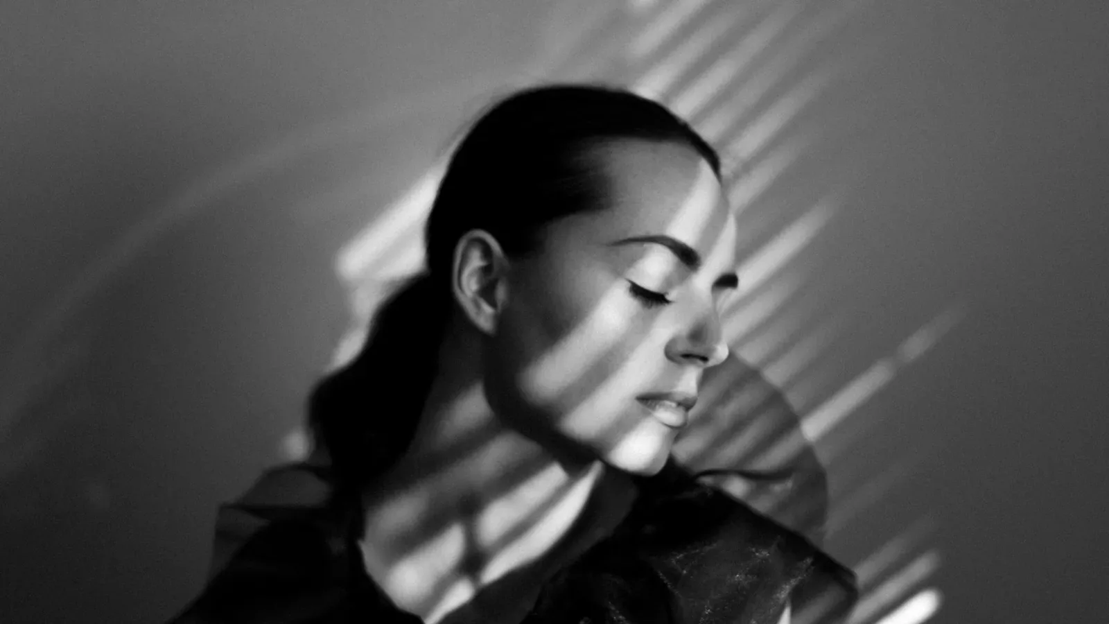 Black-and-white portrait of a woman with her eyes closed, her face crossed by bands of light and shadow.